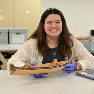 A woman wearing purple gloves holding a model watercraft from the Northwest Coast and smiling at the camera. 